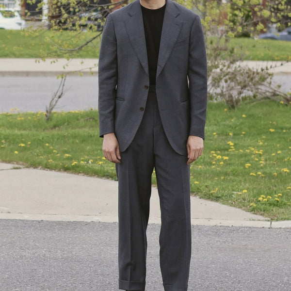 Person wearing a gray suit standing on a street with grass and flowers in the background
